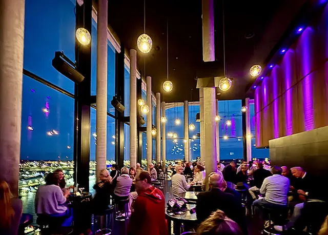 Gäste in der Lounge vor raumhohen Fenstern mit Blick auf die Lichter der Stadt